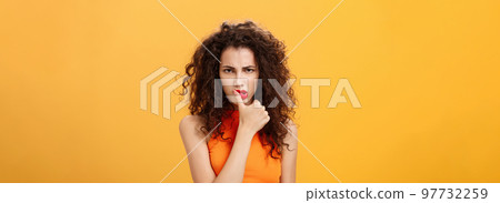 Waist-up shot of serious determined and focused displeased woman with curly hairstyle frowning biting thumb looking dangerous and angry standing daring and rebellious over orange background Waist-up shot of serious determined and focused displeased woman with curly hairstyle frowning biting thumb looking dangerous and angry standing daring and rebellious over orange background 97732259
