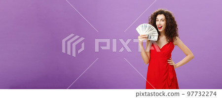 Girl knows how earn money. Successful and delighted satisfied woman in elegant red dress with curly hair holding cash near face like hand fan rejoicing from lots of cash in arms, smiling pleased 97732274
