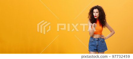 Woman seducing hot guy. Sensual and sexy daring female with curly hairstyle in stylish modern cropped top and shorts showing come here gesture inviting move closer flirting over orange background 97732459