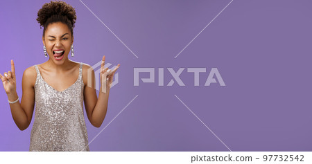 Excited good-looking african-american woman in glittering silver shiny dress having fun amusing university party prom night show tongue winking joyfully rock-n-roll heavy-metal gesture, blue wall 97732542