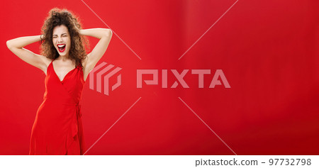Girl just wanna have fun. Crazy adult caucasian woman with curly hairstyle in elegant dress holding hands on head yelling out loud with closed eyes and intense expression over red background 97732798