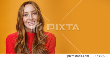 Close-up shot of tender gentle and cute redhead cutie with long natural hairstyle smiling joyfully with friendly expression posing cute and sensual against orange background, feeling happy 97733922