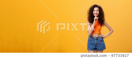 Shh I got something for you. Portrait of charming european young female with curly hairstyle in cropped top and shorts showing shush gesture with broad smile having secret preparing surprise 97733927
