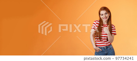 Tender cute feminine redhead lovely girl, tilt head cheerfully smiling, pointing left, showing way, give advice what purchase, advertising good product, introduce shop, orange background 97734241