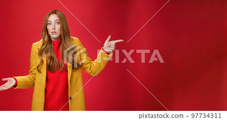 Woman missed client posing confused and unaware how happened shrugging making clueless gesture with palm as pointing right and looking questioned and uncertain at camera over red background 97734311