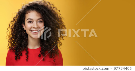 Close-up shot of pretty caucasian girl with curly hair in red dress and earrings smiling joyfully with pleased hopeful expression gazing at camera carefree, posing over yellow background 97734405