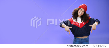 Waist-up shot of cool and stylish young woman with curly hair smiling flirty and joyful pointing down showing promotion as tilting head happily and posing over blue background in outdoor clothes 97734707