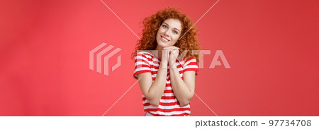 Tender feminine caring redhead curly-haired female romantic gaze camera tilt head clench palms near head silly lovely look smiling delighted cheerful dreamy sighing look with sympathy 97734708