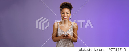 Charming elegant smiling lucky african-american woman in glittering silver dress holding smartphone grinning gladly camera texting using phone send photos after party, standing blue background Charming elegant smiling lucky african-american woman in glittering silver dress holding smartphone grinning gladly camera texting using phone send photos after party, standing blue background 97734743