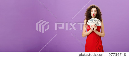 Studio shot of woman being lucky in casino winning lots of cash holding money in hands saying wow with folded lips staring impressed and excited over purple wall in evening red dress Studio shot of woman being lucky in casino winning lots of cash holding money in hands saying wow with folded lips staring impressed and excited over purple wall in evening red dress 97734748