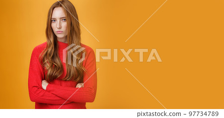 Suspicious intense and defensive ginger girl standing in passive-aggressive pose pouting and frowning looking with disbelief and disdain at camera, offended against orange background 97734789
