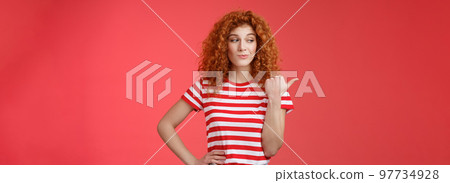 Look that stuff. Redhead curious sassy good-looking curly-haired girl summer striped t-shirt smirking gazing pointing left indicating thumb intriguing product standing red background 97734928