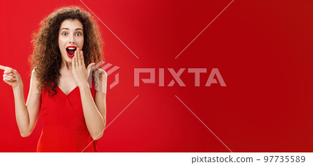 Happy surprised and delighted attractive curly-haired female in evening dress smiling amazed with opened mouth covering it with palm pointing left questioned and excited over red background 97735589