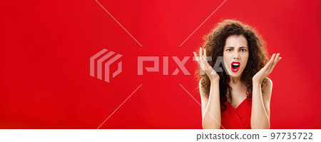 Moody displeased and angry rich woman being dissatisfied with awful chip gift raising palms in frustration and clueless gesture arguing, shouting in outrage standing in red elegant dress 97735722