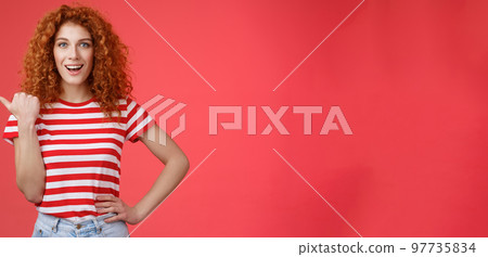 Sassy good-looking emotive happy smiling redhead european female curly hairstyle pointing thumb left grin assertive cheeky hold hand waist directing promo advertising offer red background Sassy good-looking emotive happy smiling redhead european female curly hairstyle pointing thumb left grin assertive cheeky hold hand waist directing promo advertising offer red background 97735834