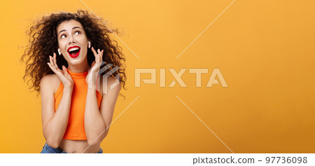Waist-up shot of attractive silly curly-haired european female in cropped top using wireless earphones touching earbuds and gazing at upper right corner delighted and carefree over orange wall Waist-up shot of attractive silly curly-haired european female in cropped top using wireless earphones touching earbuds and gazing at upper right corner delighted and carefree over orange wall 97736098