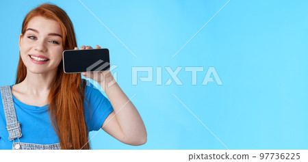 Close-up cheerful amazed cute redhead female look delighted, hold smartphone horizontal, show application, favorite phone game, stand blue background satisfied happy, grinning pleased 97736225