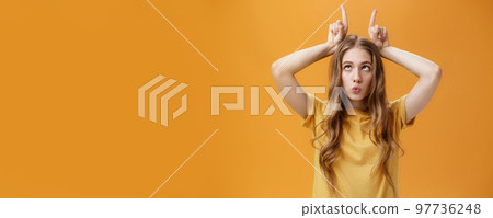 GIrl going crazy from boredom making freaky funny faces fooling around squinting holding index fingers on head like horns sticking out tongue and folding lips aping over orange background 97736248