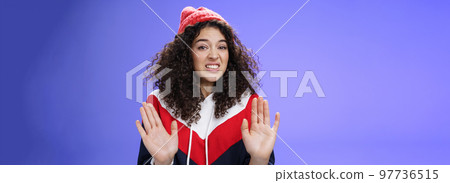 Displeased european woman with curly hair raising palms near chest and waving in refusal and rejection gesture grimacing unsatisfied and unwilling to participate in suspicious action over blue wall Displeased european woman with curly hair raising palms near chest and waving in refusal and rejection gesture grimacing unsatisfied and unwilling to participate in suspicious action over blue wall 97736515