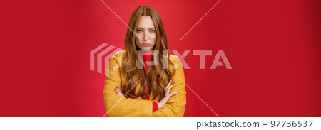 Offended and angry redhead girlfriend expressing dismay and hate as being waiting too long crossing hands over chest in defensive and aggressive pose, pouting looking with contempt over red background 97736537