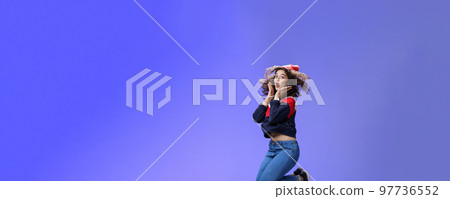 Surprised and amazed amused good-looking woman with curly hair in winter beanie and sweatshirt open mouth from joy and amazement jumping over blue background having fun feeling playful 97736552