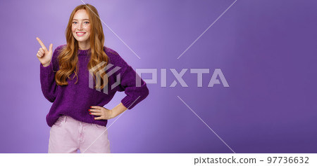 Hey click here. Portrait of friendly-looking charming and cute redhead female in purple sweater pointing at upper left corner with index finger and smiling, showing copy space, recommending, offering 97736632