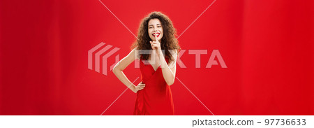 Keep it secret dear. Charming sensual and flirty adult woman with curly hairstyle smiling joyfully having mysterious plan saying shh showing shush gesture with index finger over mouth near red wall 97736633