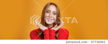 Close-up shot of sensual tender and gentle ginger girl with romance and happy gaze holding palms near pure clean skin with freckles smiling joyfully tilting head looking friendly over orange wall 97736662
