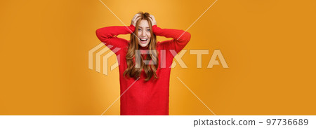 Excited and overwhelmed charming redhead woman in knitted warm red dress yelling from thrill and happiness holding hands on hair smiling broadly impressed with unexpected luck over orange wall Excited and overwhelmed charming redhead woman in knitted warm red dress yelling from thrill and happiness holding hands on hair smiling broadly impressed with unexpected luck over orange wall 97736689