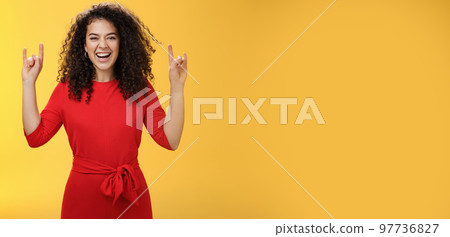 Rock n roll live. Charismatic excited young curly-haired woman in red dress feeling awesome as waiting concert of favorite band showing rock gestures with raised hands and smiling thrilled and happy Rock n roll live. Charismatic excited young curly-haired woman in red dress feeling awesome as waiting concert of favorite band showing rock gestures with raised hands and smiling thrilled and happy 97736827