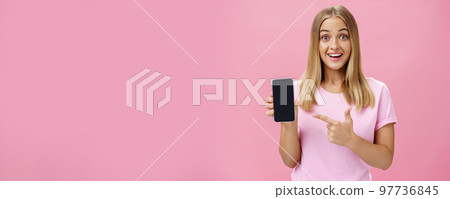 Cheerful attractive and pleasant woman promoting cool app or smartphone holding cellphone and pointing at device screen smiling amused and impressed standing over pink background 97736845