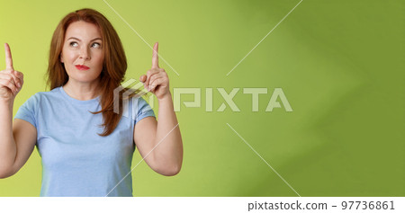 Curious thoughtful cute redhead female middle-aged ginger mother smirking thinking look aside pointing up index fingers deciding choice pondering choosing between variants green background 97736861
