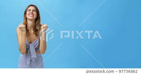 Girl triumphing raising hands in cheer from happiness and victory standing pleased and excited with closed eyes and raised head celebrating successful deal posing delighted against blue background 97736862