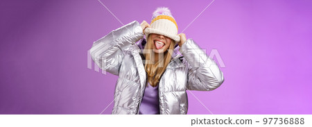 Playful carefree funny blond girl having fun fool around careless what people thing acting immature joyfully pulling hat eyes sticking tongue smiling standing silver jacket purple background 97736888