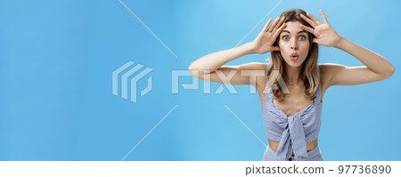 Woman looking surprised and impressed at camera after opening eyes after waiting for surprise. Emotive and playful good-looking european female holding palms near forehead folding lips curiously 97736890