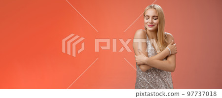 Tender feminine elegant blond young woman in stylish silver glittering dress close eyes dreamy smiling touching herself cuddle embrace hands crossed body remember heartwarming passionate feelings Tender feminine elegant blond young woman in stylish silver glittering dress close eyes dreamy smiling touching herself cuddle embrace hands crossed body remember heartwarming passionate feelings 97737018