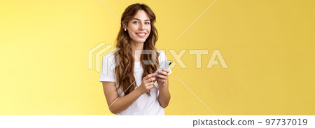 Feminine lovely curly-haired woman white t-shirt hold smartphone picked awesome new song listen music wearing wireless earphones smiling delighted camera enjoy earbuds beats yellow background Feminine lovely curly-haired woman white t-shirt hold smartphone picked awesome new song listen music wearing wireless earphones smiling delighted camera enjoy earbuds beats yellow background 97737019