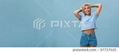 Studio shot of cute attractive and joyful stylish blond woman with tattoos on arms winking and smiling broadly holding index fingers on head making horns standing in summer outfit over blue wall Studio shot of cute attractive and joyful stylish blond woman with tattoos on arms winking and smiling broadly holding index fingers on head making horns standing in summer outfit over blue wall 97737020
