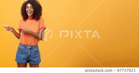 Ask him. Portrait of friendly and joyful good-looking stylish female shop assistant with curly hair and dark skin pointing left with both hands, smiling assured and entertained over orange background Ask him. Portrait of friendly and joyful good-looking stylish female shop assistant with curly hair and dark skin pointing left with both hands, smiling assured and entertained over orange background 97737021