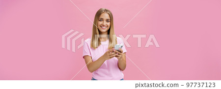 Woman calling taxy via smartphone asking friend address smiling cheerfully at camera holding phone with both hands over chest getting in touch with clients via messages standing over pink wall 97737123