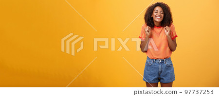 Girl eager to see lover coming from trip standing excited and thrilled with happy broad smile clenching fists grinning with closed eyes feeling joyful and amused over orange background 97737253