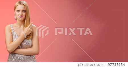 Jealous displeased angry young revengeful blond ex-girlfriend in luxurious silver shiny dress frowning pouting pointing upper right corner displeased pissed standing fed up upset red background Jealous displeased angry young revengeful blond ex-girlfriend in luxurious silver shiny dress frowning pouting pointing upper right corner displeased pissed standing fed up upset red background 97737254