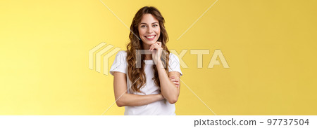 Tender feminine enthusiastic charming european woman curly long hairstyle laughing silly gaze grinning joyfully touch chin intrigued standing curious interested listening amused yellow background 97737504