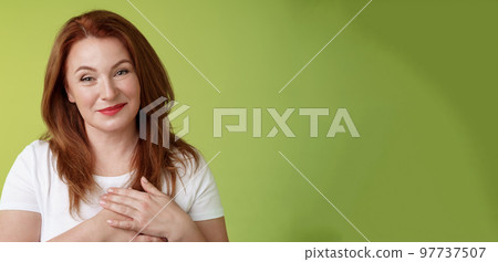 Lovely pleased tender redhead middle-aged ginger woman press hands heart feel touched grateful appreciate sincere heartwarming moment admire thankfully grin look camera glad green background 97737507