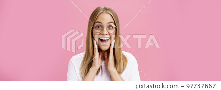 Woman liking new glasses picked in store feeling happy finding right frame touching cheeks from happiness smiling cheerfully at camera wearing transparent eyewear and t-shirt over pink wall Woman liking new glasses picked in store feeling happy finding right frame touching cheeks from happiness smiling cheerfully at camera wearing transparent eyewear and t-shirt over pink wall 97737667