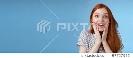 Amused happy dreamy young excited redhead female fan looking fascinated camera smiling admiration delighted touch face thrilled express emotional cheerful surprise, blue background 97737925
