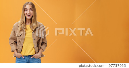 Charismatic tanned woman in corduroy jacket and yellow t-shirt ready for chilly autumn walk with friends smiling joyfully gazing entertained at camera holding hand in pockets casually over orange wall 97737933