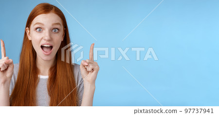 Surprised happy enthusiastic young redhead woman reacting impressed upper hanging promo pointing up index fingers drop jaw amused look thrilled camera telling about awesome offer, blue background 97737941