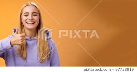 Amused joyful optimistic young excited blond european female model in hoodie smiling broadly close eyes having fun show approval gesture thumbs-up like awesome trip vacation idea, orange background Amused joyful optimistic young excited blond european female model in hoodie smiling broadly close eyes having fun show approval gesture thumbs-up like awesome trip vacation idea, orange background 97738019