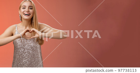Adorable happy charming lovely blond european girl in silver dress express love positivity show heart gesture confess sympathy passionate feelings towards boyfriend smiling broadly, red background 97738113